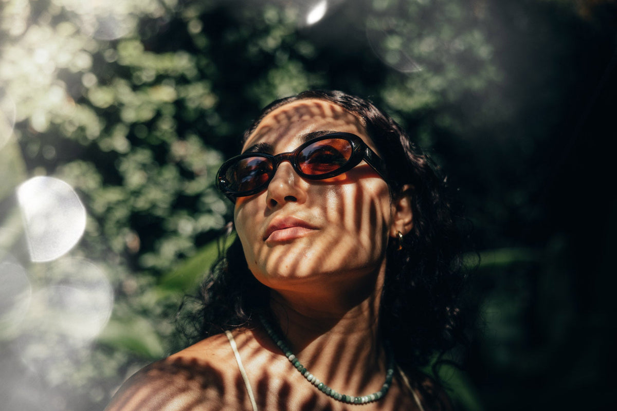 Woman wearing sunglasses in a leafy outdoor setting with dappled shadow patterns across her face, edited with ATMOS Pro Suite light and glass overlay for a bokeh and shadow effect